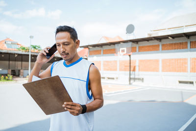 Man talking on phone while holding clipboard at basketball court