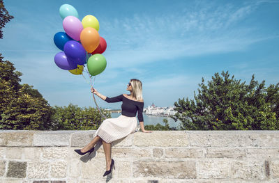 Woman holding balloons while sitting on retaining wall against blue sky