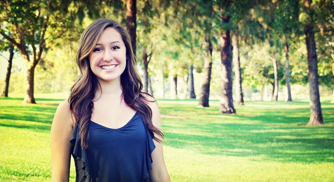 young adult, looking at camera, portrait, person, young women, tree, long hair, front view, smiling, lifestyles, grass, leisure activity, casual clothing, park - man made space, focus on foreground, standing, beauty