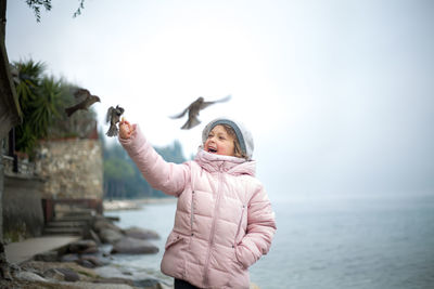 Girl feeding birds while standing by sea against sky