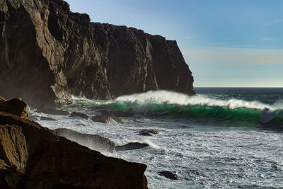 Waves splashing on rocks at shore against sky