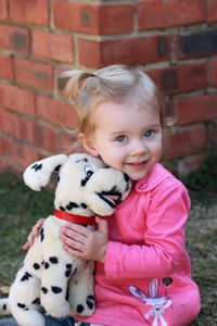 Portrait of cute girl with stuffed toy sitting in yard against brick wall