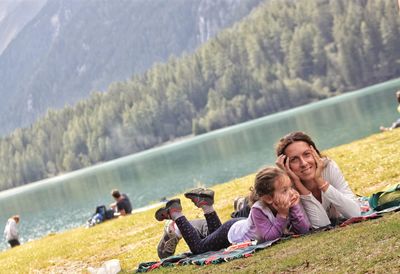 Portrait of mother lying with daughter at lakeshore against trees