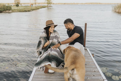 Smiling woman in blanket sitting by man and dog on pier against lake