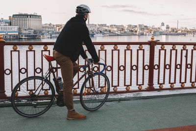 Side view of male cyclist with bicycle standing on bridge over river against sky in city