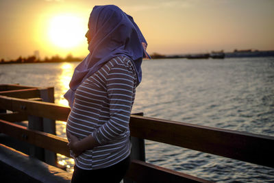 Midsection of woman standing by railing against sea during sunset