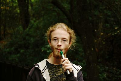 Portrait of young man smoking electronic cigarette while standing against trees