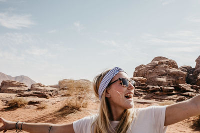 Happy young woman with arms outstretched standing at desert against sky