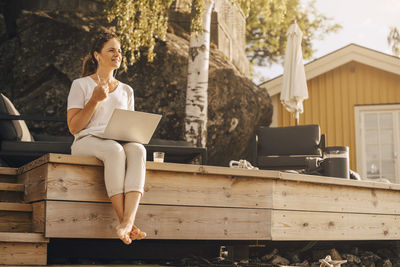 Full length of woman talking on headphones while using laptop at holiday villa