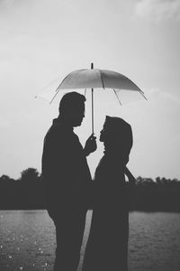 Silhouette man holding umbrella standing by lake against sky