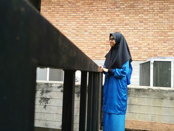 Smiling young woman standing by railing against brick wall