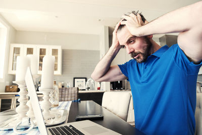 Frustrated young man with head in hands using laptop at home