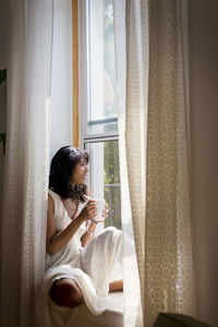 Thoughtful woman looking away while sitting on window sill at home