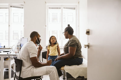 Male doctor with stethoscope looking at woman talking to girl in medical clinic