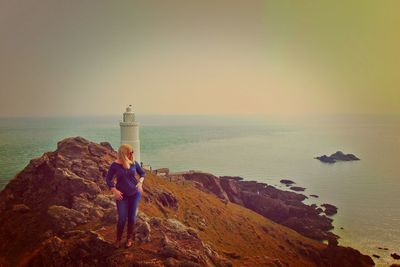 High angle view of woman standing on cliff against lighthouse by sea during sunset