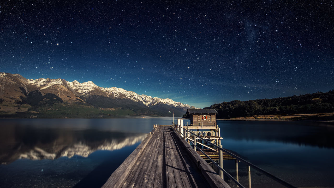 Pier over lake by snowcapped mountains against star field