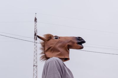 Low angle view of person wearing horse mask against electricity pylon