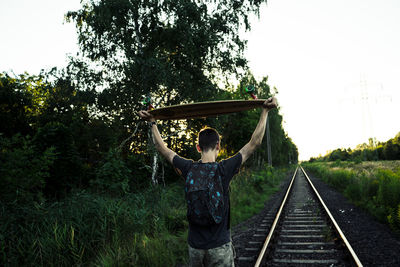 Rear view of man carrying skateboard while standing on railroad track