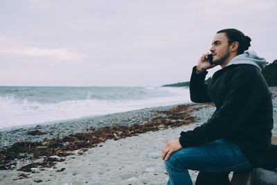 Side view of young man using mobile phone while sitting at beach