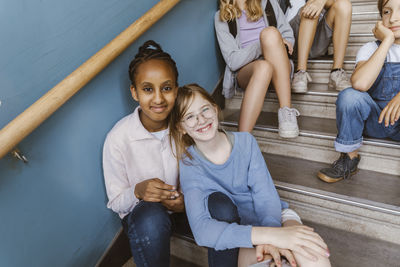 Portrait of smiling multiracial female friends sitting on staircase in school building