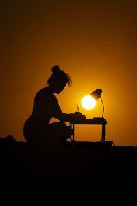 Optical illusion of silhouette woman writing while sitting at table against orange sky during sunset