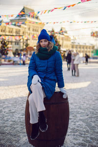 Woman in blue jacket and scarf and white gloves sit  barrel in winter, square in moscow on christmas