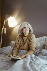 Thoughtful senior woman with book sitting on bed at home