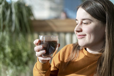 Smiling young caucasian woman holding glass cup of coffee on background of green plants, drinking