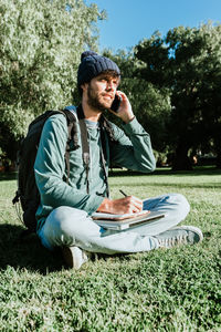Full length of man talking on phone while sitting on grass outdoors