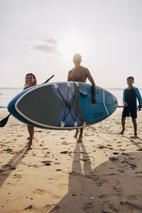 Man carrying surfboard while walking with son and daughter on sand at beach against sky