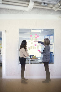Side view of female business professionals discussing over adhesive notes stuck on glass in office