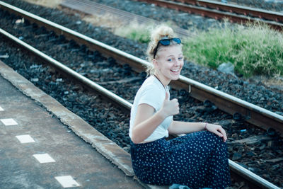 Side view of cheerful young woman gesturing thumbs up while sitting by railroad track