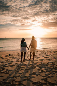 Rear view of friends standing on beach against sky during sunset