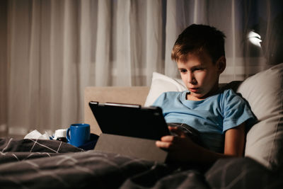 Little boy using tablet while laying in bed in living room. medicines and thermometer nearby. 