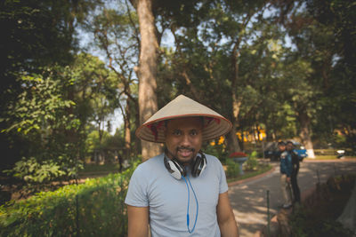 Portrait of man wearing asian style conical hat and headphones at park