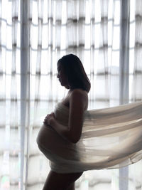 Side view of pregnant woman standing against window