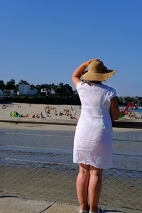 Rear view of woman wearing hat standing on sidewalk against clear sky during sunny day
