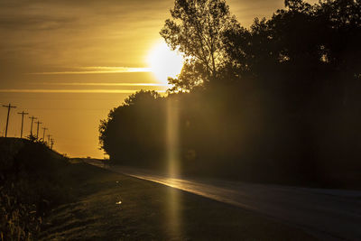 Silhouette trees by road against sky during sunset