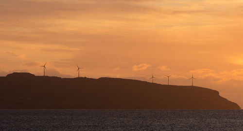 Silhouette windmill by sea against orange sky
