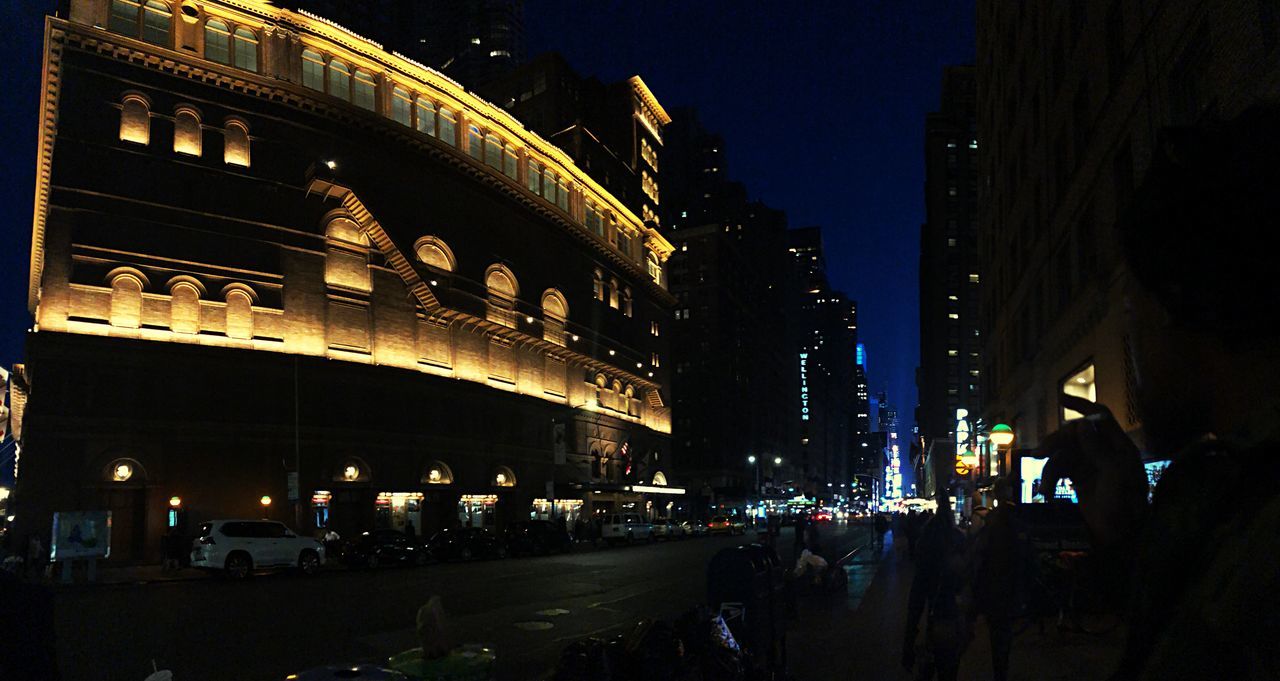 PEOPLE WALKING ON ILLUMINATED ROAD AMIDST BUILDINGS AT NIGHT