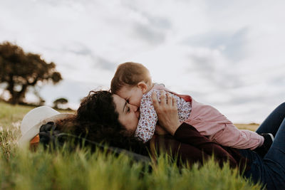 Mother kissing daughter while lying on lawn against sky