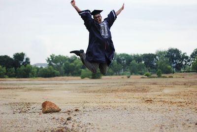 Full length of cheerful man in graduation gown jumping on field