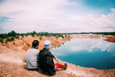 Rear view of man and woman sitting by lake against cloudy sky