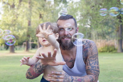 Smiling bearded man holding son playing with bubbles in park