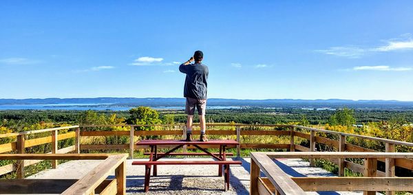 Rear view of man looking at landscape while standing on bench against sky