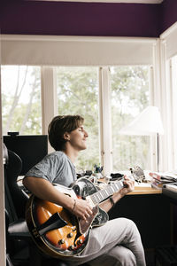 Young man with eyes closed playing guitar while sitting at home