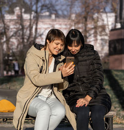 Two young women with a smile sit on a bench in sunny park and look into one smartphone