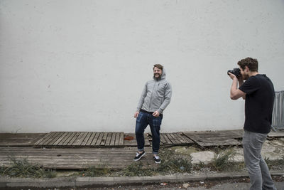 Man photographing cheerful friend against wall on footpath