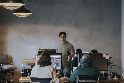 Young male hacker discussing with male and female colleagues working on computer in startup company