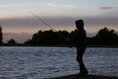 Man fishing on lake against sky during sunset